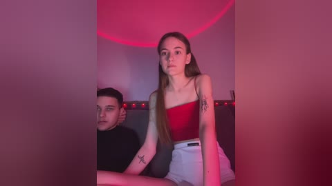 Media: A video of a young woman with long hair in a red tank top and high-waisted shorts, sitting with a tattooed man in a dimly lit, red-lit room.