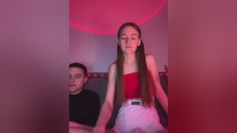 Media: Video of a young, fair-skinned woman with long brown hair in a red top, sitting on a bed, next to a man in a black shirt. The room is dimly lit with red ambient lighting.