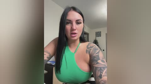 Video of a pale-skinned woman with long black hair, wearing a green halter top, showcasing detailed tattoos on her arms, standing indoors with a beige wall and a framed art piece in the background.
