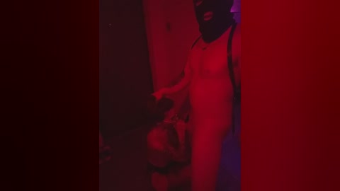 Video of a dark-skinned man with a muscular build, wearing a black mask, standing in a dimly lit room with red lighting. He holds a person's head, partially obscured by shadow.