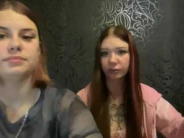 Video of two teenage girls in a dimly lit room with a dark, patterned wallpaper. The girl on the left has fair skin, straight brown hair, and wears a grey sweater. The girl on the right has fair skin, long brown hair with red streaks, and wears a pink jacket over a floral top.