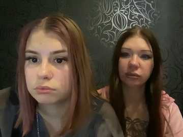 Video of two young women with fair skin and straight, long brown hair. One woman has a neutral expression, wearing a dark jacket. The other, with a neutral expression, wears a pink jacket. Both have dark eyes and light makeup. Background has ornate wallpaper and a chandelier.