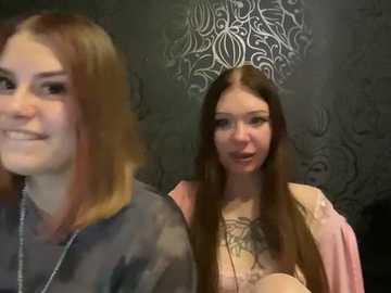 Video of two young women, one with long auburn hair and the other with long brown hair, smiling, in a room with a gray, ornate wallpaper and a silver chandelier.