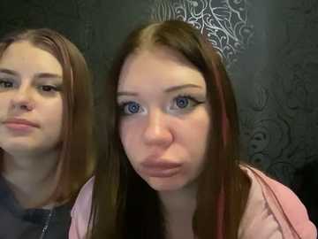 Media: Video of two young Caucasian women with fair skin and long straight brown hair. One has a serious expression, while the other puckers her lips in a silly manner. Dark, patterned wallpaper in the background.