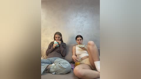 Media: Video of two young women with long hair, one in a gray hoodie, the other in a white crop top, eating food on a beige sofa against a textured wall.