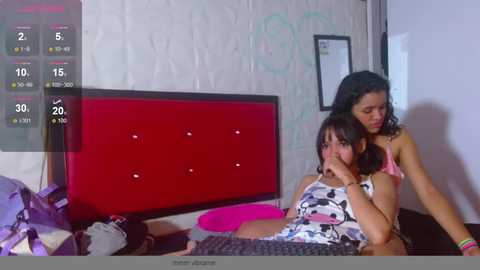 Video of two women, one with a large chest, lying on a bed with a red headboard; the other, with a smaller bust, sits behind her. The room has a cluttered, casual atmosphere.