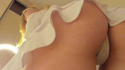 Media: Video of a blonde woman's lower body in a white skirt, revealing beige panties, taken from a low angle emphasizing her buttocks. Background shows a mirror and beige walls.