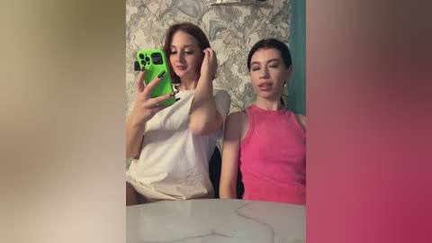 Media: Video of two women taking selfies in a bathroom. One wears a white top, the other a pink tank top. Wallpaper and sink visible.