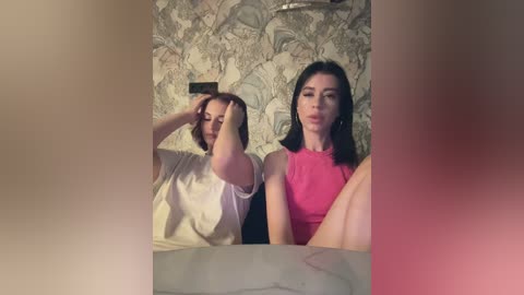 Media: Video of two women in a dimly lit room with a floral wallpaper background. One woman, in a white shirt, covers her face with her hand, while the other, in a pink dress, sits next to her.