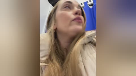Video of a Caucasian woman with long blonde hair, wearing a beige jacket, looking up thoughtfully. Background features a blue curtain and blurred, light-colored walls, suggesting a bathroom setting.
