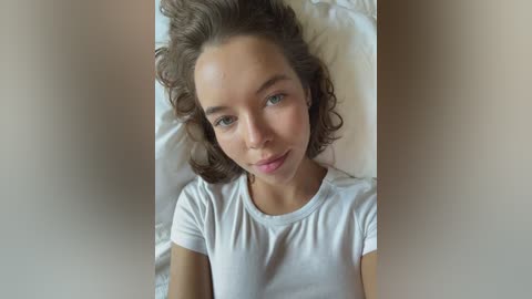 Media: A video of a young woman with curly brown hair, fair skin, and blue eyes, lying on a white pillow, wearing a white t-shirt, in a serene indoor setting.