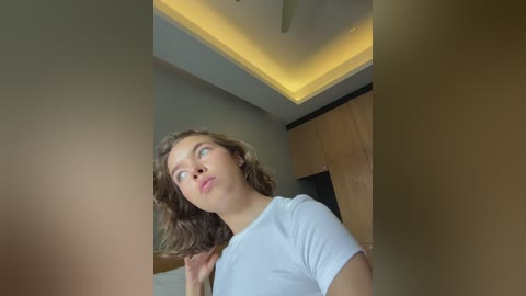 Media: Video of a young woman with wavy brown hair, wearing a white t-shirt, standing indoors in a modern room with a ceiling light and wooden wall.