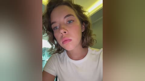 Media: A close-up video of a young woman with curly brown hair and light skin, wearing a white T-shirt. The background features a blurred indoor setting with soft, ambient lighting.