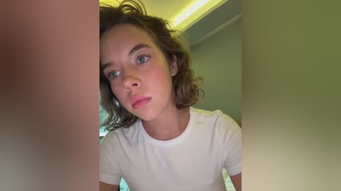 Media: Video of a young Caucasian woman with short, wavy brown hair, light skin, and blue eyes, wearing a white t-shirt, taken indoors with a soft, ambient light.