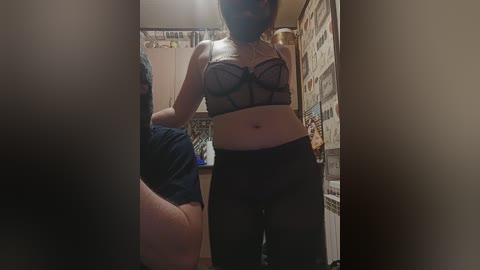 Video of a slender, light-skinned woman with a black mask, wearing a sheer black bra and black pants, standing in a dimly lit bathroom with a cluttered background.