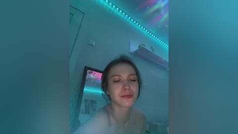A video of a young woman with fair skin, brown hair, and closed eyes, wearing a white top, in a dimly lit room with teal LED lights, and a TV in the background.