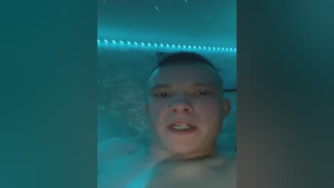 Video of a young, shirtless Asian man with short dark hair, visible braces, and a slightly open mouth, standing in a dimly lit, teal-lit, steamy bathroom.