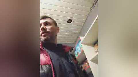 Media: Video of a bearded man with short dark hair, wearing a black leather jacket, standing in a narrow, dimly-lit hallway with white tiled ceiling and beige walls.