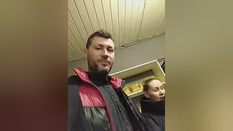 Media: A video shows a man in a red and black puffy jacket, standing indoors, with a woman in a black jacket behind him. The room has a beige ceiling and shelves with yellow items.