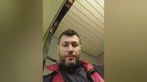 Media: Video of a middle-aged man with short dark hair, beard, and mustache, wearing a black jacket with a red collar. He stands in a room with beige walls and a wooden ceiling.