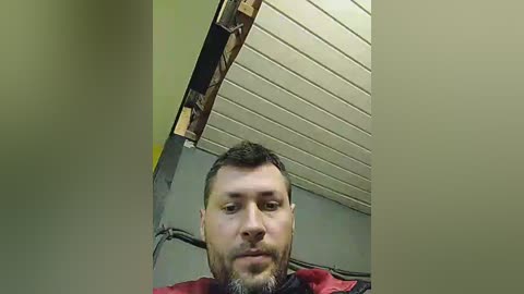 Media: Video of a middle-aged man with short dark hair, a beard, and mustache, wearing a red jacket, standing in a room with exposed wooden ceiling beams and gray walls.