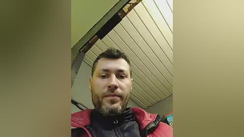 Media: A video of a man with a short beard, wearing a red and black jacket, standing in a room with a beige ceiling and exposed metal beams.