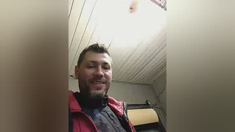 Media: A video of a smiling man with short brown hair and a beard, wearing a red jacket and black shirt, standing in a small, dimly-lit room with white paneling and a fluorescent light fixture.