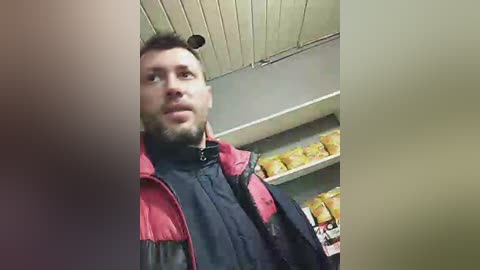 Media: Video of a man with short dark hair and beard, wearing a red and black puffer jacket, standing in a grocery store aisle with shelves of packaged food.