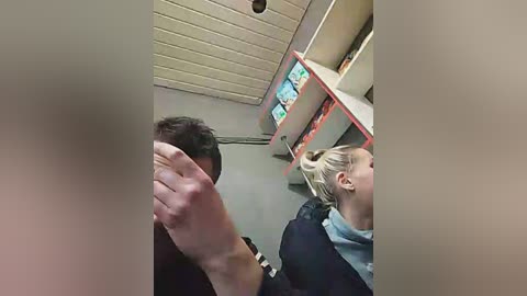 Media: Video of a woman with blonde hair in a gray coat and scarf, and a man with dark hair in a black jacket, standing in a library with shelves of books and computer screens.