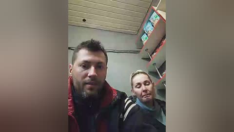 Media: Video of a man with short brown hair and a beard in a red jacket, standing next to a woman with blonde hair in a black jacket. They are in a small, dimly lit room with a TV screen displaying a video game in the background.