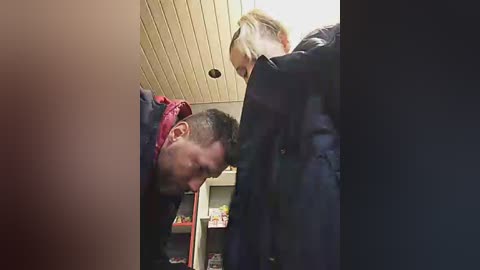Media: Video of a man with a red shirt and a woman with blonde hair, both bending over to examine something on a shelf in a room with a light-colored ceiling.