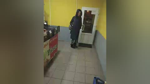 Media: Video of a woman in a dark coat and boots, standing near a vending machine with a red and white \"Pepsi\" sign, in a dimly lit hallway with yellow and blue walls.