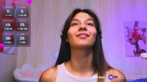 Video of a smiling, light-skinned woman with long black hair in a white tank top, standing in a room with a white wallpaper and abstract art.