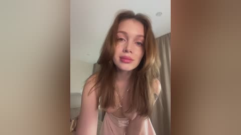 Video of a young Caucasian woman with light skin and long, wavy brown hair, wearing a pale pink satin robe, standing indoors against a beige wall and partially visible curtains.