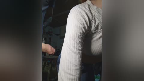 Video of a man's back in a grey ribbed sweater, holding a large, erect penis. The background is a dimly lit room with various objects and furniture blurred.