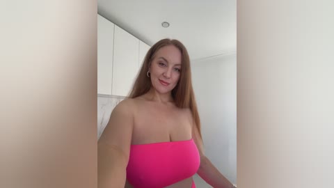 Media: Video of a fair-skinned, long-haired, smiling Caucasian woman in a bright pink strapless top, standing in a minimalist, white-walled kitchen.