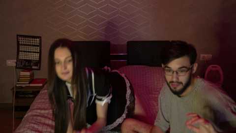 Media: A video of a woman in a maid outfit, bending over a man in a sweater on a bed with patterned bedding. Background shows a wall-mounted chessboard and a pink heart-shaped pillow.