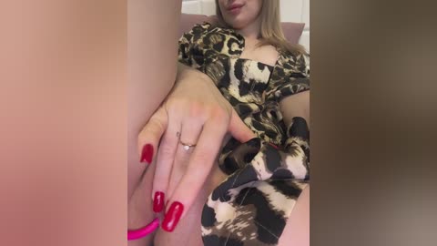 A video of a fair-skinned woman with blonde hair, wearing leopard-print pajamas and red nail polish, seated and holding a pink vibrator.