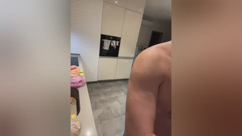Media: Video of a nude, muscular man from the side, standing in a modern, minimalist kitchen with white cabinets, stainless steel appliances, and gray tile floors.