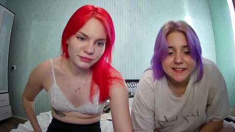 Media: Video of two young women with vibrant red and purple hair, wearing casual clothes, seated on a bed against a pastel green wall.