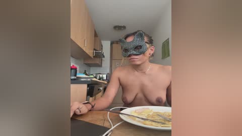 Video of a topless woman with medium-sized breasts wearing a black and white mask, sitting at a kitchen table with a plate of food. The kitchen background includes cabinets, a stove, and a microwave.