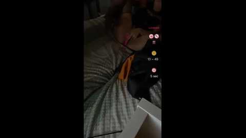 A video shows a person with medium skin tone, wearing a black hat, lying on a bed with gray sheets, holding a smartphone, surrounded by social media icons.