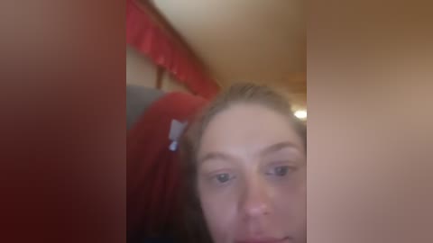 A blurry video of a young woman with light skin and brown hair, partially visible, in a red jacket, taken indoors with warm lighting.
