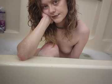 Video of a fair-skinned woman with curly brown hair, partially submerged in a white bathtub, leaning forward with her chin resting on her hand.