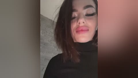 Video of a young woman with fair skin and dark hair, wearing black makeup, with her eyes closed, against a gray brick wall.