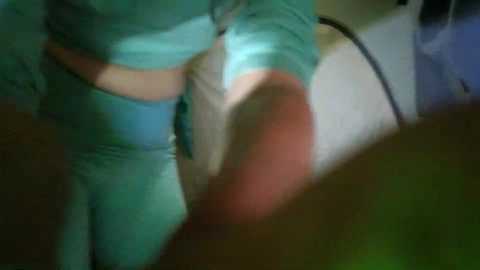 A blurry video of a person in teal scrubs bending over, partially visible in a dimly lit room with a white cabinet and dark background.