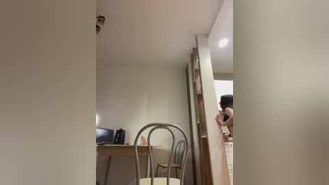 Video of a small, sparsely furnished room with beige walls, a desk, chair, and a mirror. A woman in a black top leans against the mirror.