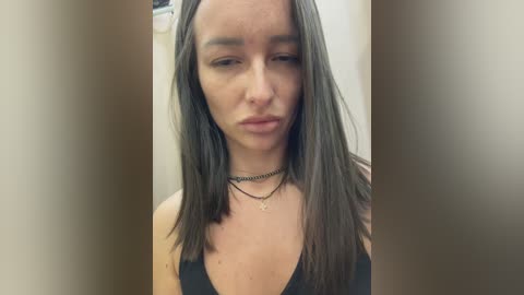 Media: Video of a young woman with long, straight brown hair, wearing a black tank top, and multiple thin, black necklaces. She has a serious expression, with a neutral background.