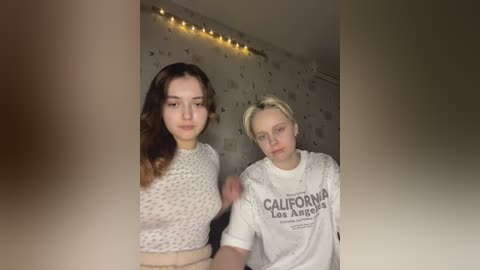 Media: Video of two young women in a dimly lit room; one with long brown hair, wearing a white polka-dot top, the other with short blonde hair, wearing a \"California Love Los Angeles\" t-shirt.