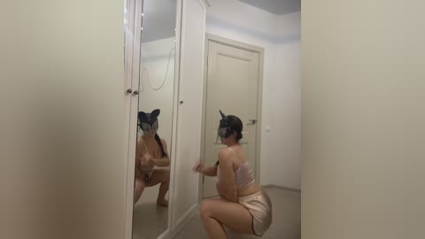 Video of a woman with black hair wearing a headband, white top, and shorts, crouching in front of a large mirror, adjusting her reflection.